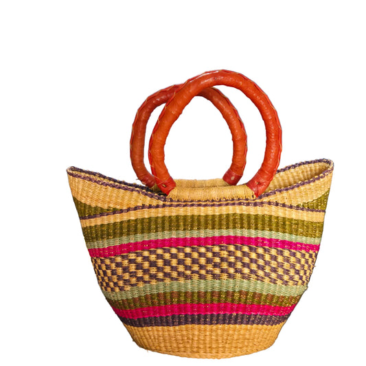 A woven straw tote bag with a striped pattern in olive green, brown, pink, and beige, featuring double curved brown handles, photographed in an office setting against a blue wall.