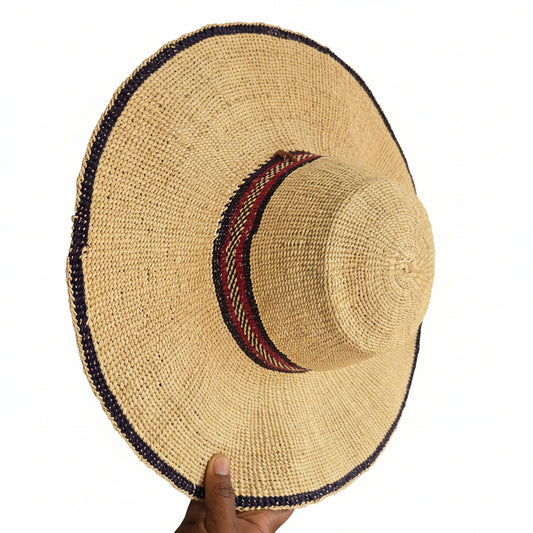 A woven straw sun hat with a wide brim, featuring a red and black patterned band around the crown and a dark blue trim along the brim edge, held by a hand.