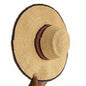 A woven straw sun hat with a wide brim, featuring a red and black patterned band around the crown and a dark blue trim along the brim edge, held by a hand.