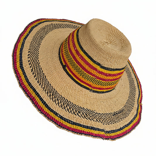 A natural straw sun hat with a wide brim, featuring woven stripes of black, red, yellow, and beige around the crown and brim edge, shown being worn on a person's head.