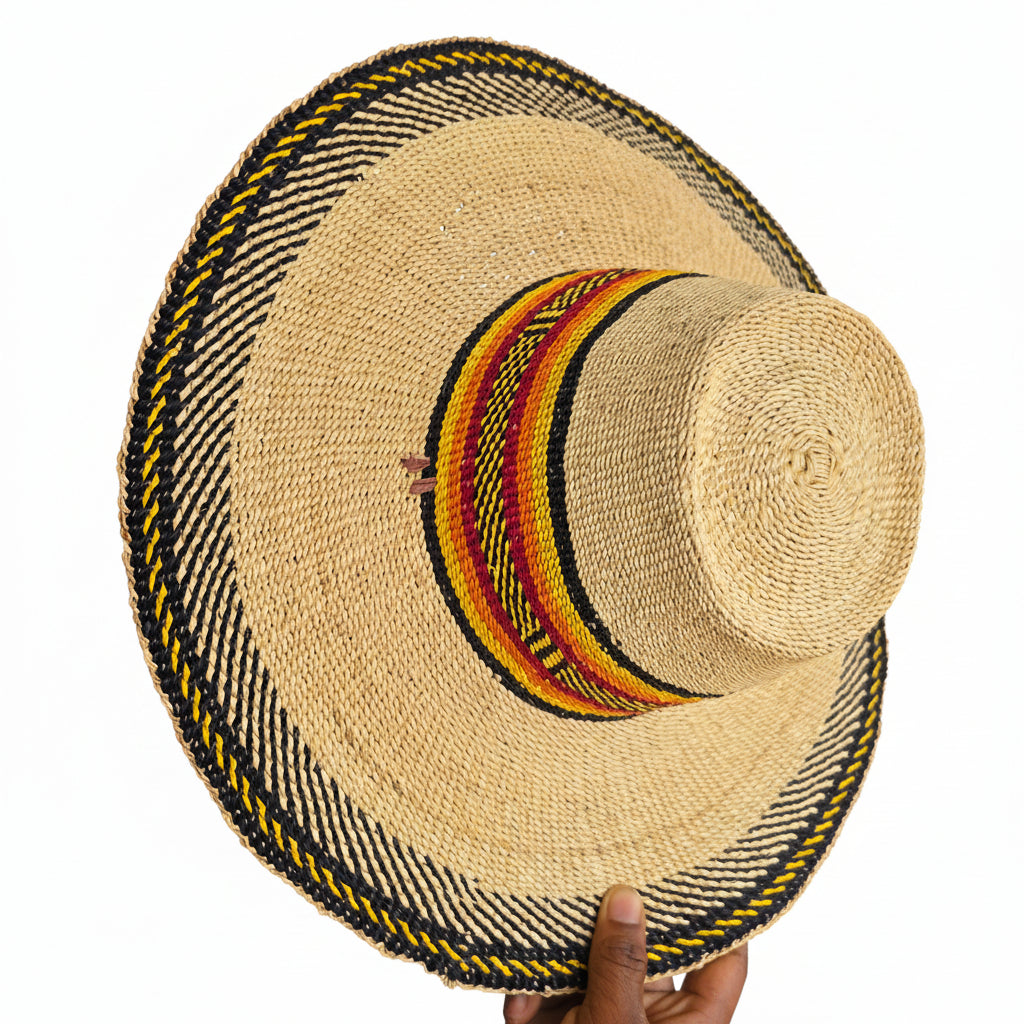 A natural straw boater-style hat with a wide brim, featuring a black, yellow, and red patterned band around the crown and a braided black and yellow trim along the brim edge. A hand is holding the brim on the right side.