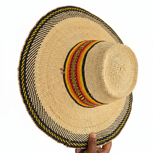 A natural straw boater-style hat with a wide brim, featuring a black, yellow, and red patterned band around the crown and a braided black and yellow trim along the brim edge. A hand is holding the brim on the right side.