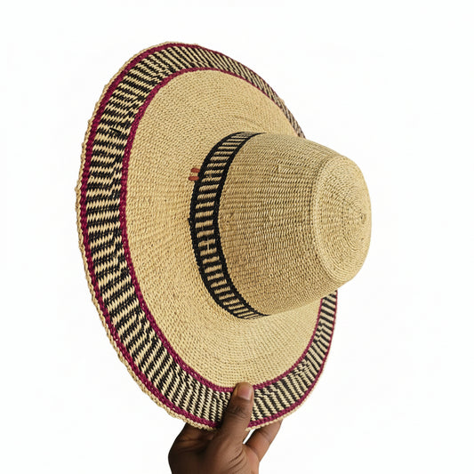 A natural straw sun hat with a wide brim, featuring a black woven band and a red and black patterned trim, held by a hand on a gray surface.