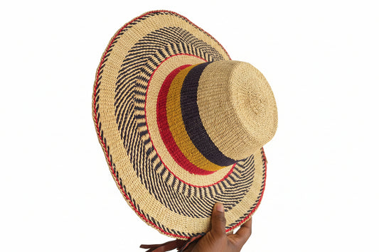 A woven straw sun hat with a wide brim, featuring a natural beige base with concentric bands of navy blue, yellow, and red, held by a hand on a dark gray shelf.