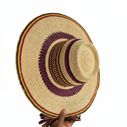 A hand holds a wide-brimmed straw hat with a natural beige base and woven bands in red, yellow, black, and white around the crown and brim, set against a workshop background.