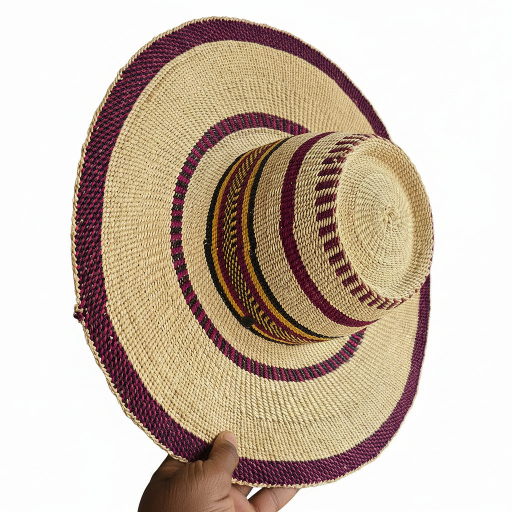 A hand-woven straw hat with a wide brim, featuring a natural beige straw body decorated with dark purple braided bands and a patterned band in red, yellow, and black. A hand is holding the brim on the right side.