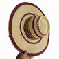 A hand-woven straw hat with a wide brim, featuring a natural beige straw body decorated with dark purple braided bands and a patterned band in red, yellow, and black. A hand is holding the brim on the right side.
