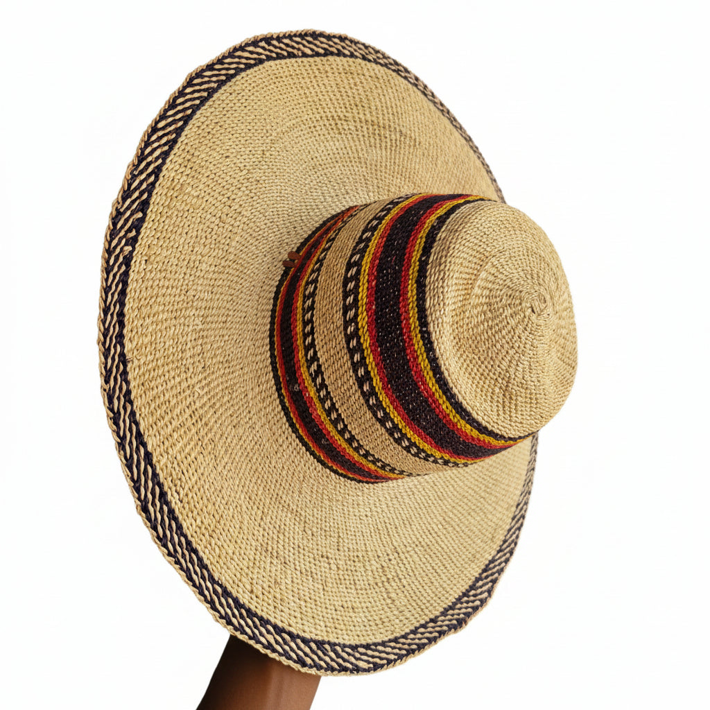 A wide-brimmed straw hat with a decorated band of red, yellow, black, and white stripes on the crown and a braided trim around the brim, held by a hand on a dark surface.