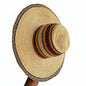 A wide-brimmed straw hat with a decorated band of red, yellow, black, and white stripes on the crown and a braided trim around the brim, held by a hand on a dark surface.