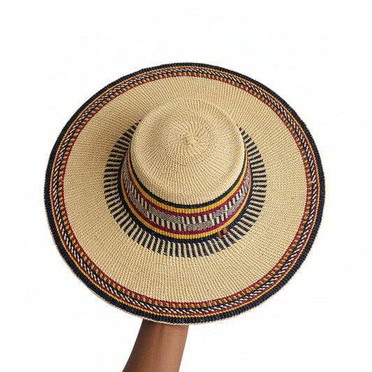 A hand-woven straw sun hat with a wide brim, featuring a natural beige straw body and a decorative band with black, red, yellow, and white woven patterns. A hand is holding the brim on the right side.