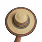 A hand-woven straw sun hat with a wide brim, featuring a natural beige straw body and a decorative band with black, red, yellow, and white woven patterns. A hand is holding the brim on the right side.