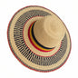 A woven straw sun hat with a wide brim, featuring black, red, and yellow stripes on the crown band and brim edge, held by a hand on a dark counter.