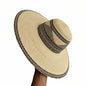 A woven straw sun hat with a wide brim and a black patterned band, shown resting on a shelf in what appears to be a workshop or storage area.