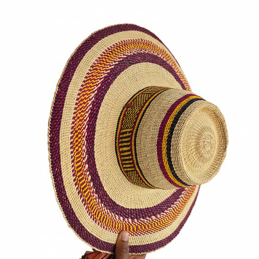 A woven straw boater-style hat with a wide brim, featuring concentric bands of purple, orange, yellow, and black woven patterns around a natural beige straw base. A hand is holding the brim on the right side in an indoor workshop or store setting.
