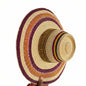 A woven straw boater-style hat with a wide brim, featuring concentric bands of purple, orange, yellow, and black woven patterns around a natural beige straw base. A hand is holding the brim on the right side in an indoor workshop or store setting.