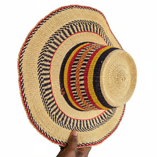 A hand-woven straw boater hat with a natural beige base and colorful striped bands in navy, red, yellow, and black, held by a hand against a workshop background.