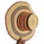 A hand-woven straw boater hat with a natural beige base and colorful striped bands in navy, red, yellow, and black, held by a hand against a workshop background.