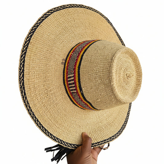 A natural straw wide-brim hat with a black braided trim and a colorful woven band in red, yellow, and black around the crown, shown on a workbench with a hand holding the brim edge.