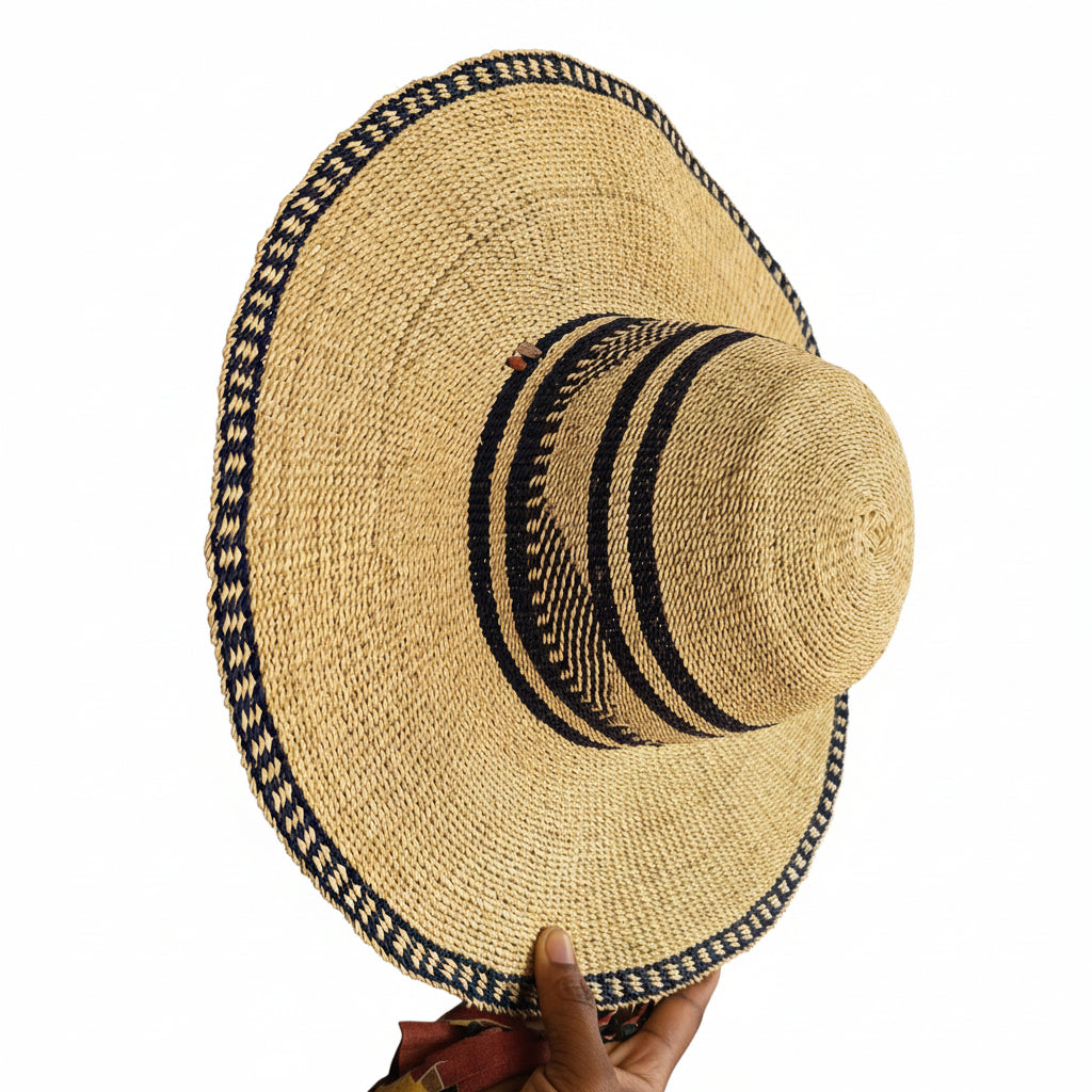 A natural straw sun hat with a wide brim, featuring black and beige woven stripes on the crown and a matching patterned trim along the brim's edge, held by a hand on the right side.