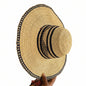 A natural straw sun hat with a wide brim, featuring black and beige woven stripes on the crown and a matching patterned trim along the brim's edge, held by a hand on the right side.