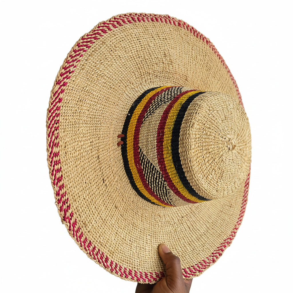 A hand holds a large woven straw sombrero-style hat against a doorway. The hat has a natural beige straw body with a red-and-white braided trim around the brim and crown, and a striped band in black, yellow, red, and white around the center of the crown.