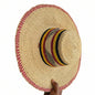 A hand holds a large woven straw sombrero-style hat against a doorway. The hat has a natural beige straw body with a red-and-white braided trim around the brim and crown, and a striped band in black, yellow, red, and white around the center of the crown.