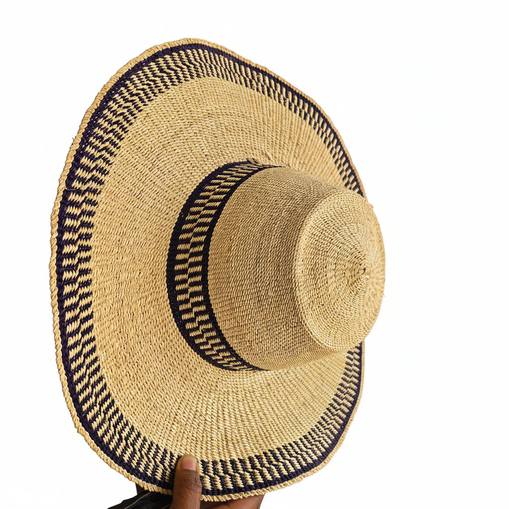 A natural straw wide-brim hat with a dark navy blue patterned band, held by a hand in a workshop setting.