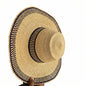 A natural straw wide-brim hat with a dark navy blue patterned band, held by a hand in a workshop setting.