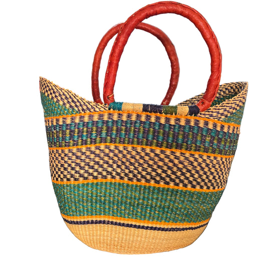 A woven straw tote bag with a striped pattern in green, black, yellow, and natural beige, featuring two curved reddish-brown leather handles, shown on a desk.