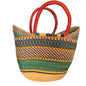 A woven straw tote bag with a striped pattern in green, black, yellow, and natural beige, featuring two curved reddish-brown leather handles, shown on a desk.