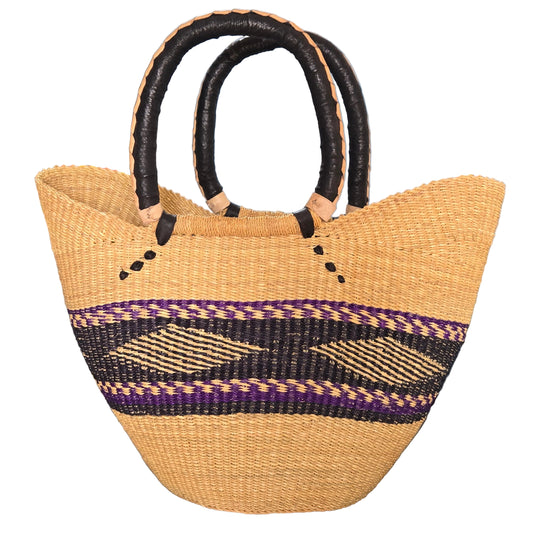 A woven straw tote bag with black faux-leather handles, featuring a purple and black geometric pattern band across the middle, set on a dark table against a blue wall.