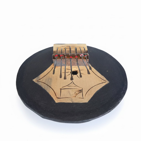 A hand holding a small wooden kalimba thumb piano. The instrument has a black-painted round body with a natural wood center featuring engraved geometric patterns and a metal tuning bridge with colorful decorative beads.