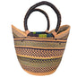 A hand-woven basket bag with dark brown twisted handles, featuring vertical stripes of black, beige, and olive green on a woven body, photographed on a desk.