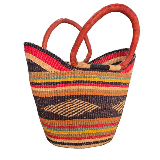 A hand-woven basket bag with terracotta-colored handles, featuring vertical stripes and diamond patterns in red, yellow, orange, green, and dark brown on a beige base, photographed on a desk.