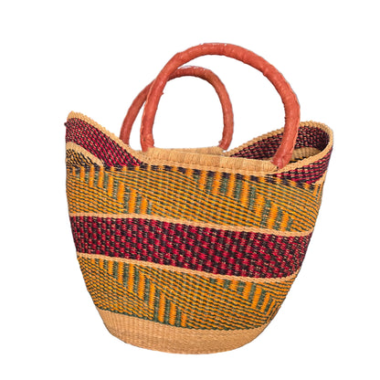 A woven basket bag with two curved handles, featuring a natural beige base, a vertical dark red stripe, and horizontal stripes of yellow, green, and red. The bag is photographed on a desk with a blue binder and office items in the background.