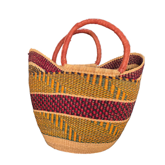 A woven basket bag with two curved handles, featuring a natural beige base, a vertical dark red stripe, and horizontal stripes of yellow, green, and red. The bag is photographed on a desk with a blue binder and office items in the background.