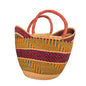 A woven basket bag with two curved handles, featuring a natural beige base, a vertical dark red stripe, and horizontal stripes of yellow, green, and red. The bag is photographed on a desk with a blue binder and office items in the background.