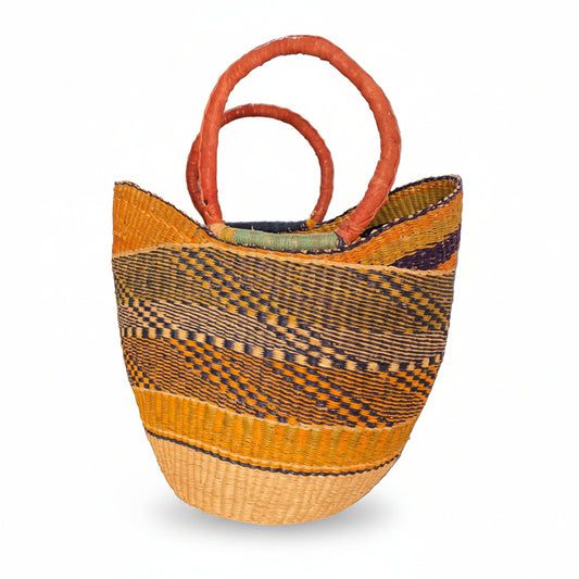 A large woven basket bag with a geometric pattern in yellow, brown, and black, featuring two thick orange-brown woven handles, photographed in a cluttered indoor setting.