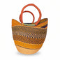 A large woven basket bag with a geometric pattern in yellow, brown, and black, featuring two thick orange-brown woven handles, photographed in a cluttered indoor setting.