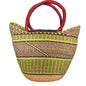 A woven straw tote bag with dark brown and lime green geometric stripes and thick red handles, photographed in a cluttered indoor setting.