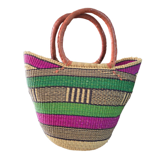 A woven straw tote bag with vertical stripes in green, purple, beige, and brown, featuring two large curved brown handles, photographed in a shop setting.